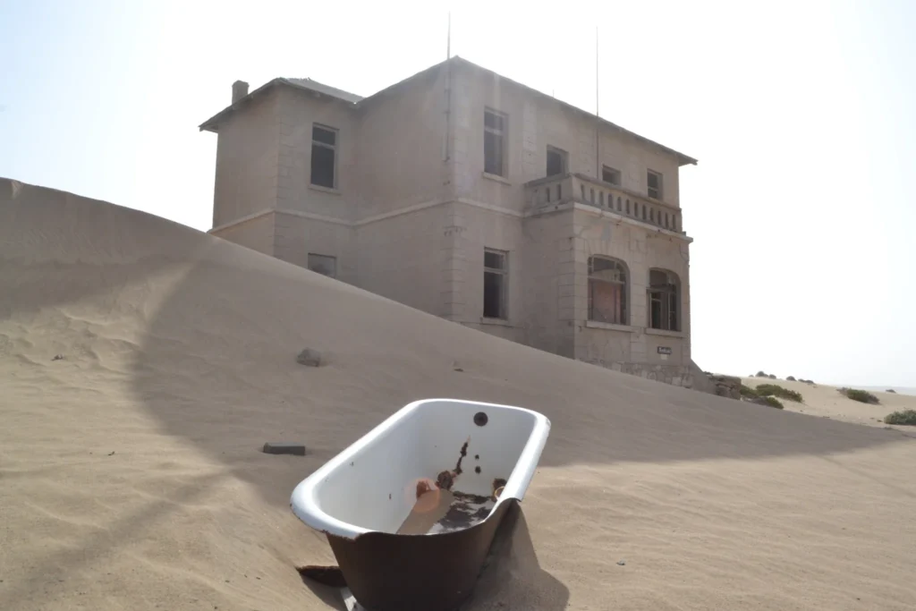 Old bathtub standing in desert sand in front of an abandoned Kolmanskop house