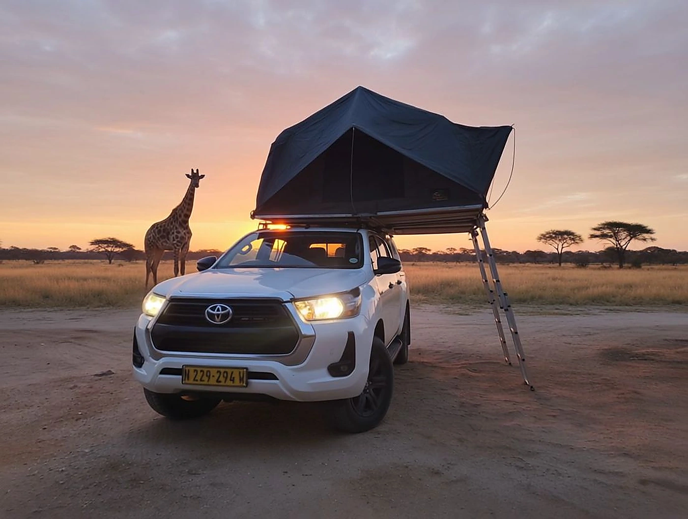 Toyota Hilux con tenda da tetto accanto a una giraffa al tramonto in Namibia